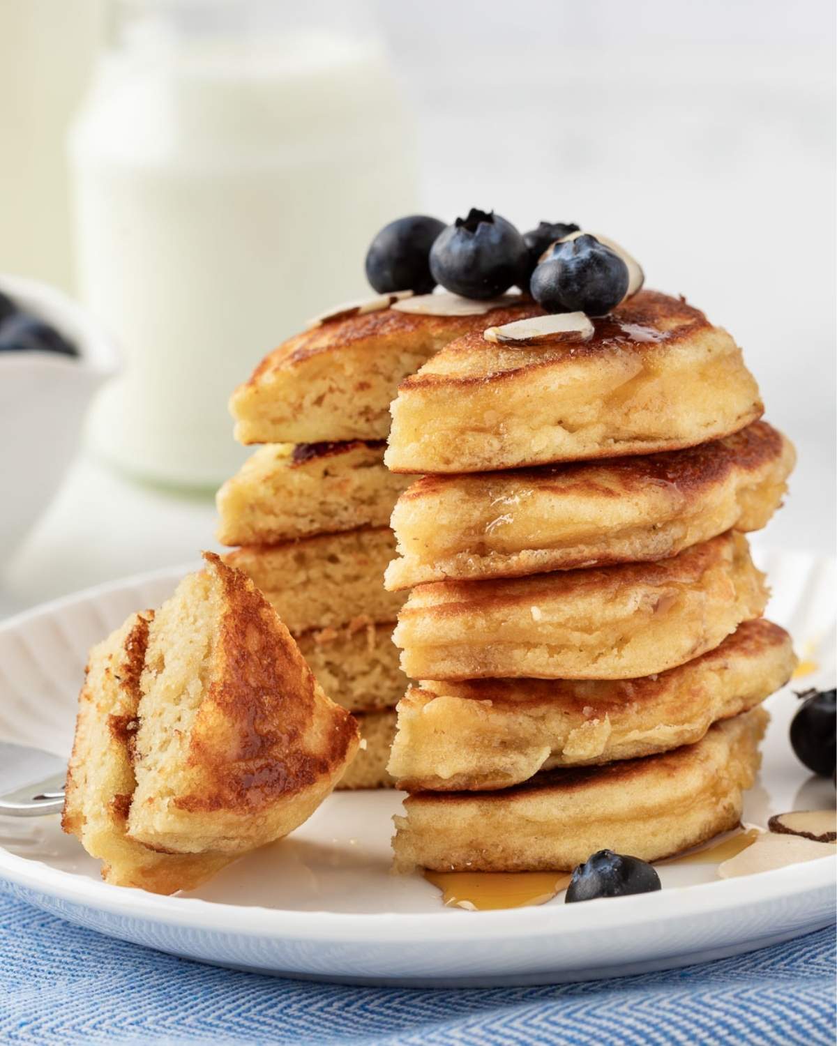 Front view of a stack of almond flour pancakes that's been sliced to show their soft and fluffy texture on a plate topped with blueberries and some almond slivers with a little maple syrup drizzled on top.