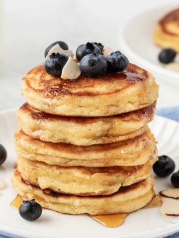 Front view of a fluffy stack of almond flour pancakes on a plate topped with blueberries and some almond slivers with a little maple syrup drizzled on top.