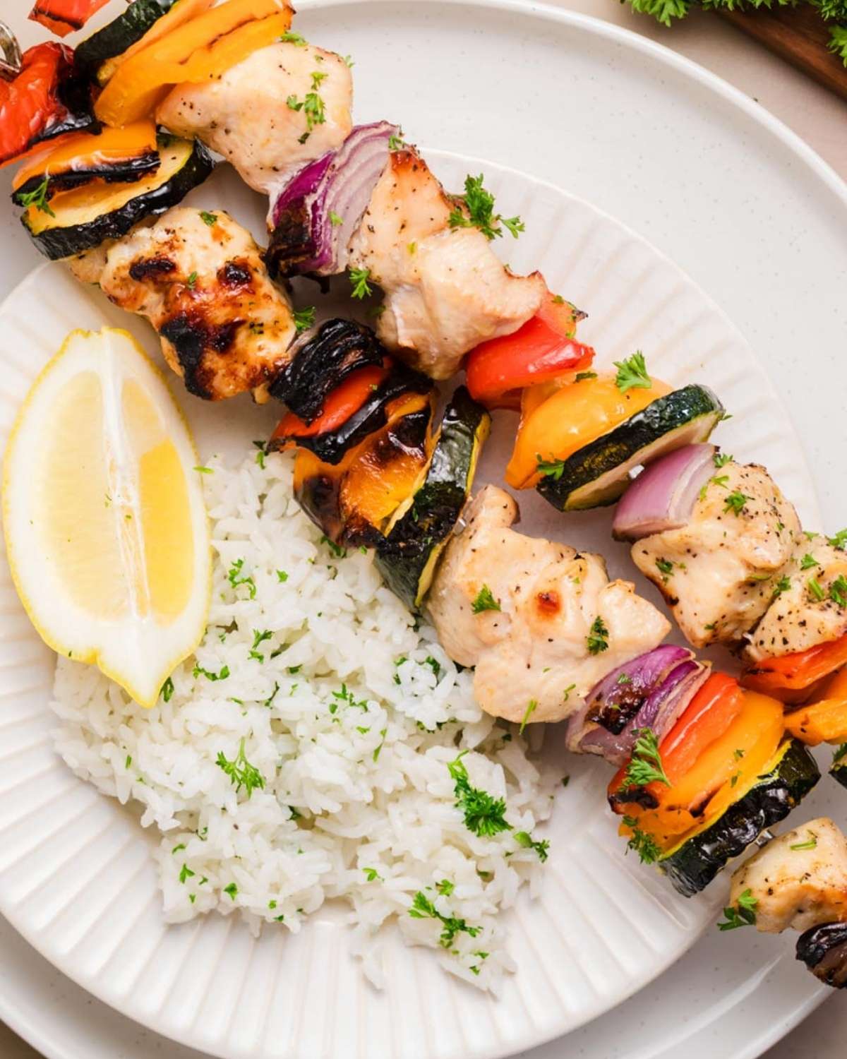 overhead view of two grilled chicken kabobs on a white plate served with herbed rice and a lemon wedge. Each skewer has chicken and veggies (bell peppers, onions, and zucchini) threaded through.