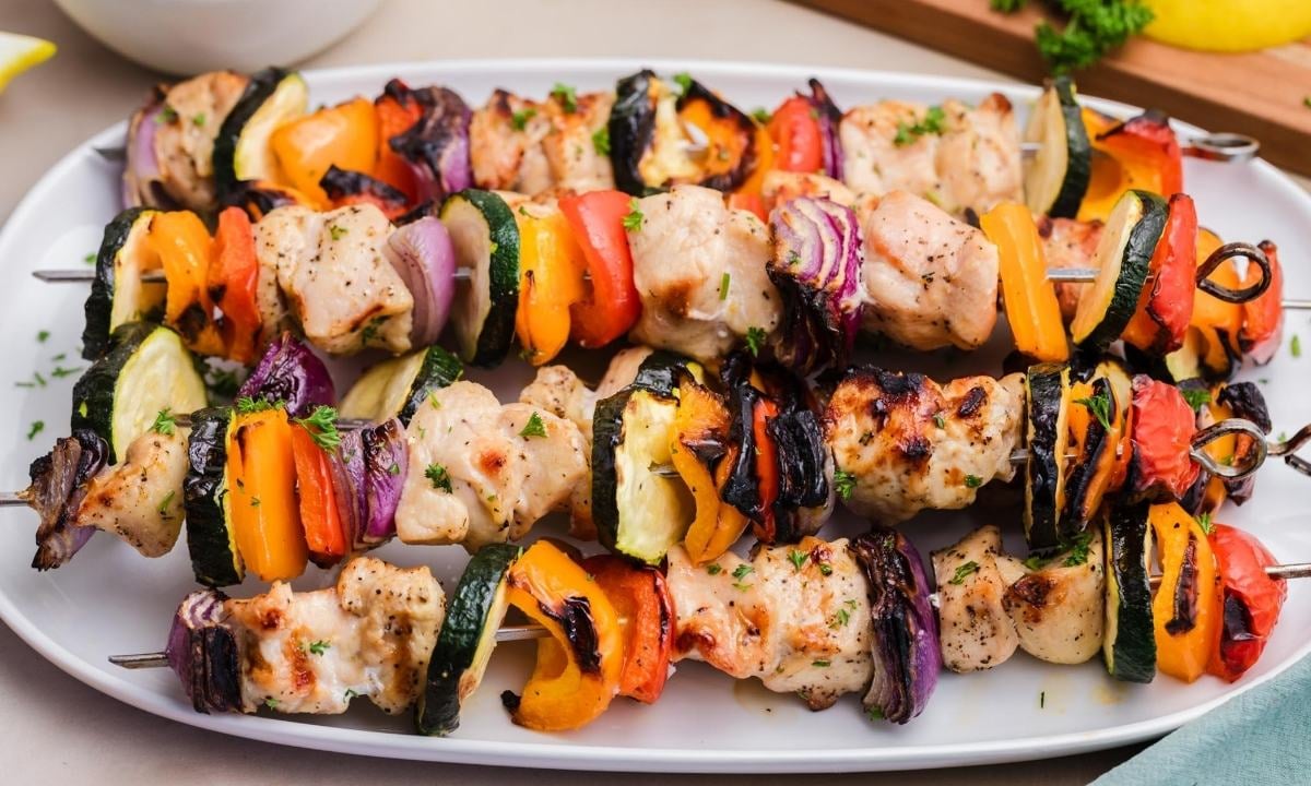 front view of stacked grilled chicken kabobs on a serving platter. Each skewer has chicken and veggies (bell peppers, onions, and zucchini) threaded through.