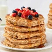 fluffy stack of oatmeal pancakes on a plate topped with blueberries and diced strawberries with a drizzle of pancake syrup
