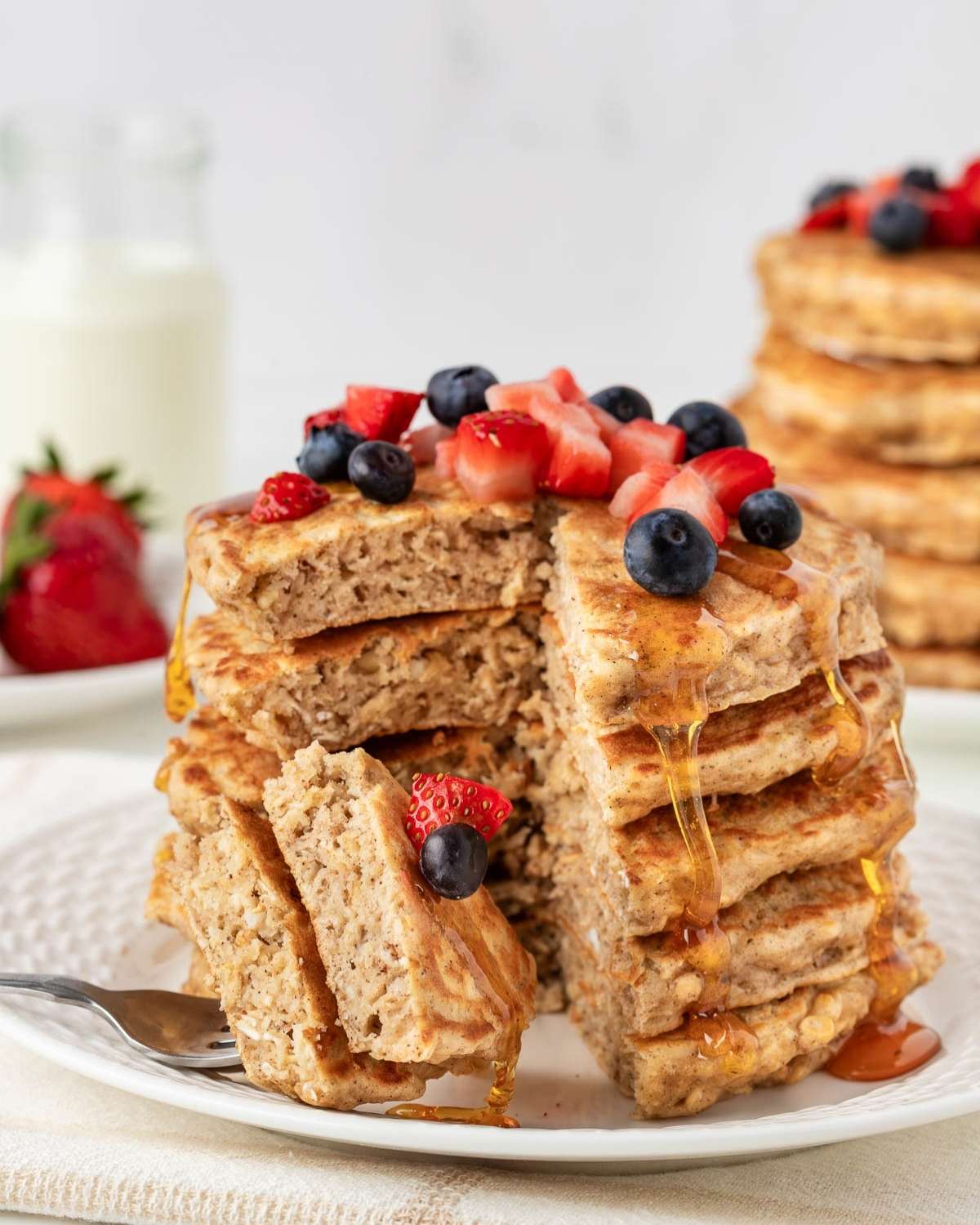fluffy stack of oatmeal pancakes on a plate with a slice on a fork, topped with blueberries and diced strawberries with a drizzle of pancake syrup