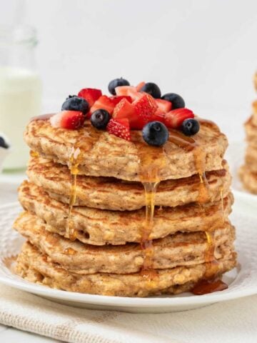 fluffy stack of oatmeal pancakes on a plate topped with blueberries and diced strawberries with a drizzle of pancake syrup