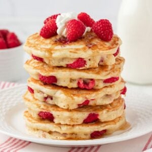 Stack of fluffy raspberry pancakes on a plate topped with fresh raspberries, whipped cream, and a drizzle of syrup.