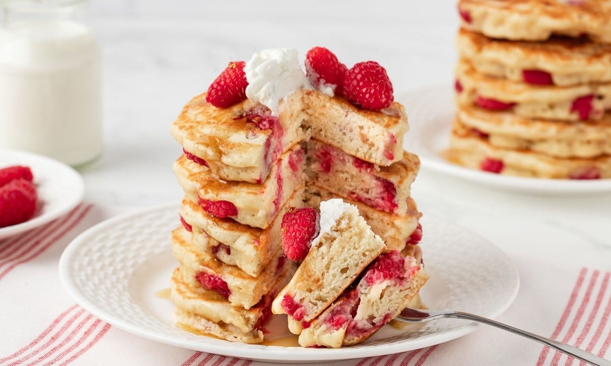Stack of fluffy raspberry pancakes on a plate topped with fresh raspberries, whipped cream, and a drizzle of syrup with the pancakes sliced and on a fork to show their fluffy texture.