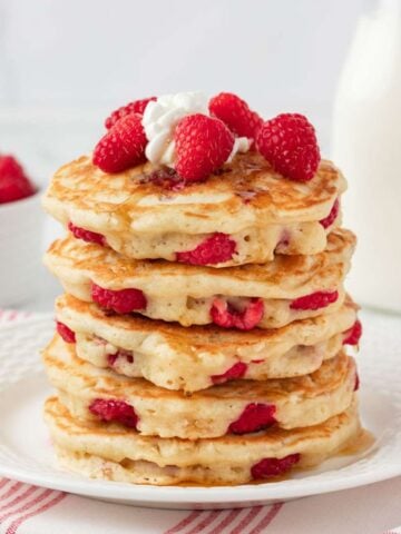 Stack of fluffy raspberry pancakes on a plate topped with fresh raspberries, whipped cream, and a drizzle of syrup.