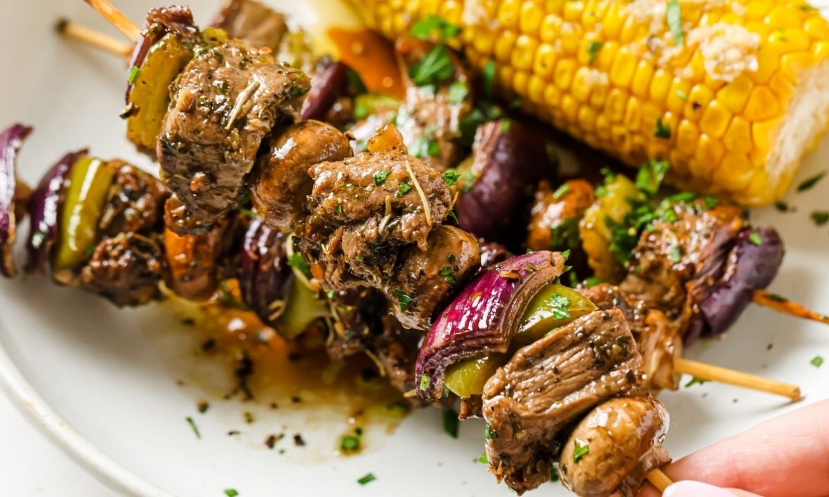 two grilled beef kabobs on a plate next to a corn on the cob