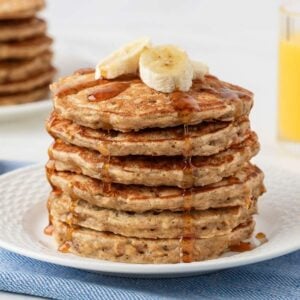 stack of banana oatmeal pancakes on a plate topped with sliced bananas and a drizzle of pancake syrup