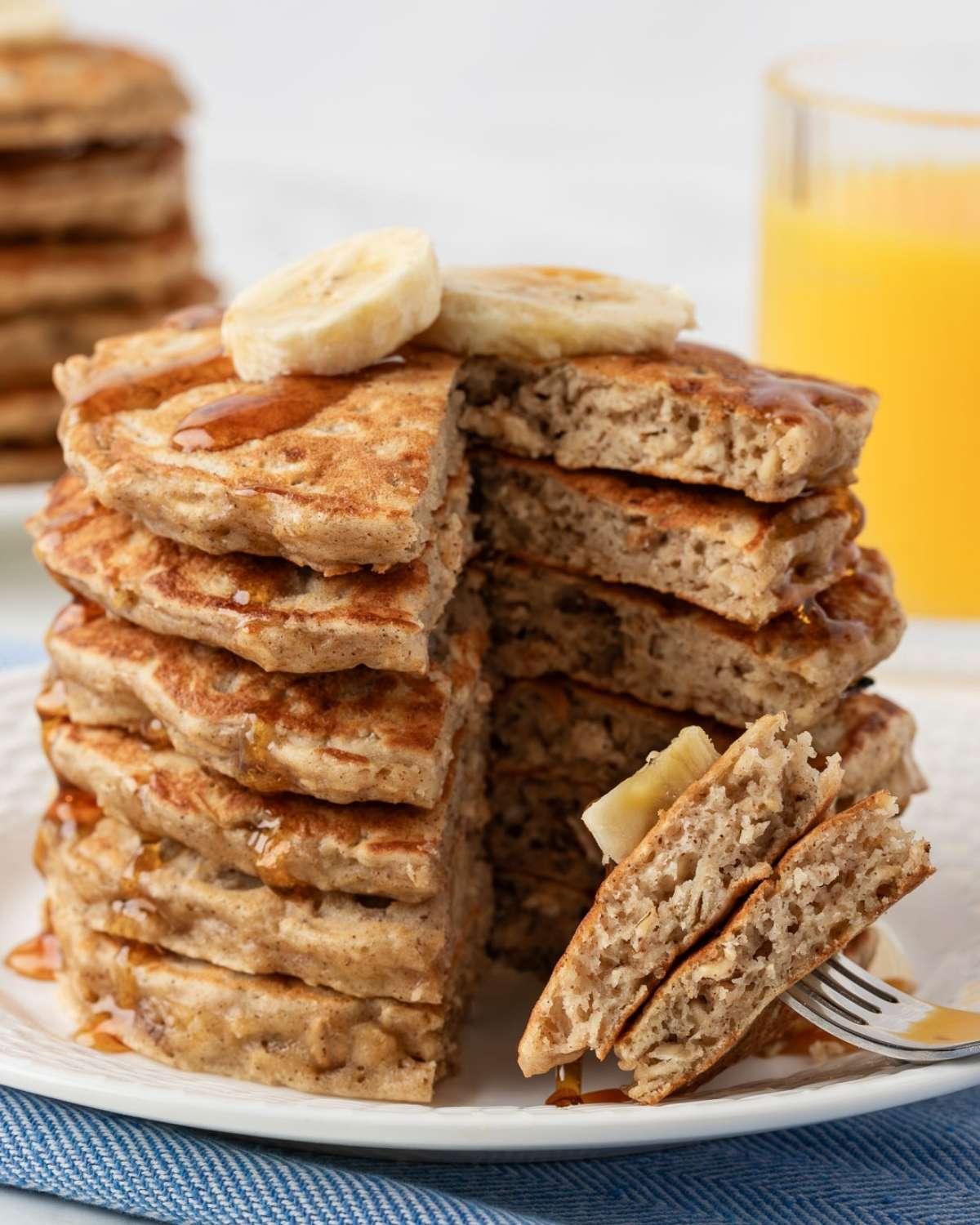 sliced stack of banana oatmeal pancakes on a fork to show hearty texture with stack on a plate topped with sliced bananas and a drizzle of pancake syrup