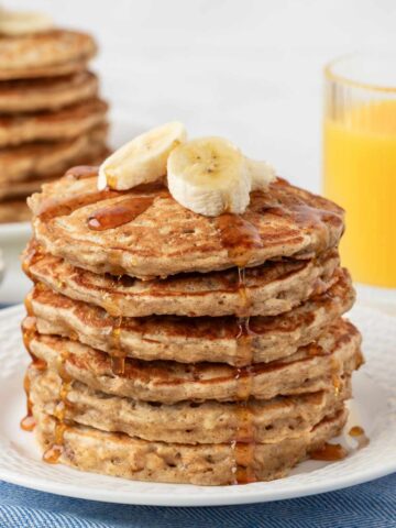 stack of banana oatmeal pancakes on a plate topped with sliced bananas and a drizzle of pancake syrup