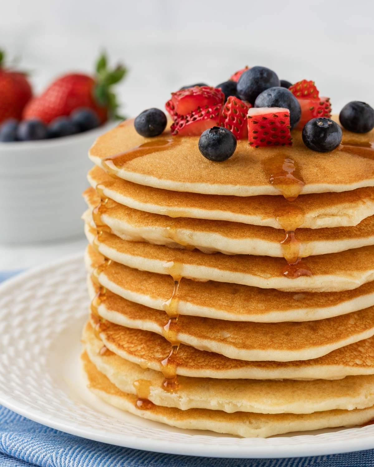 fluffy stack of pancakes made with rice flour on a plate. topped with diced strawberries and blueberries and drizzled with pancake syrup