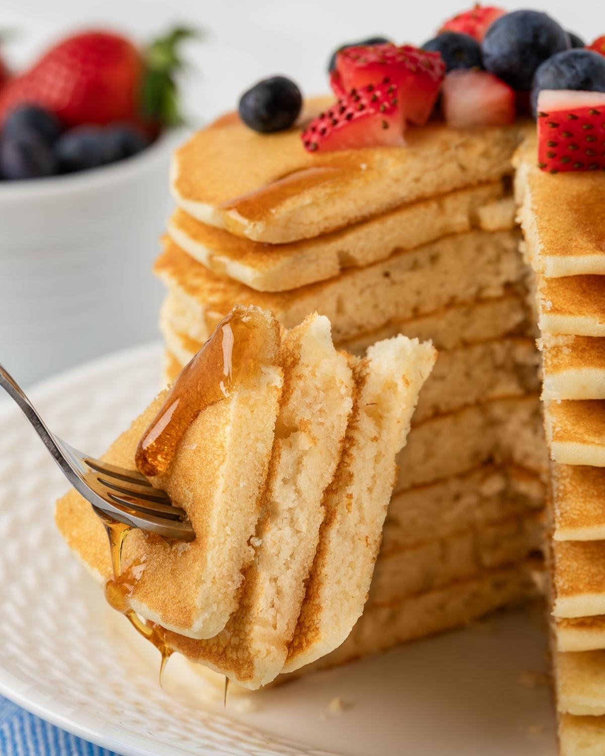 a slice of a couple of fluffy rice flour pancakes on a fork from the stack that's on the plate. they're topped with diced strawberries and blueberries and drizzled with pancake syrup