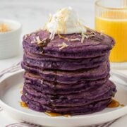 stack of purple ube pancakes on a plate topped with coconut whipped cream and toasted shredded coconut