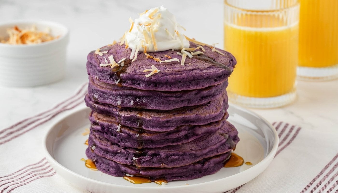 stack of purple ube pancakes on a plate topped with coconut whipped cream and toasted shredded coconut