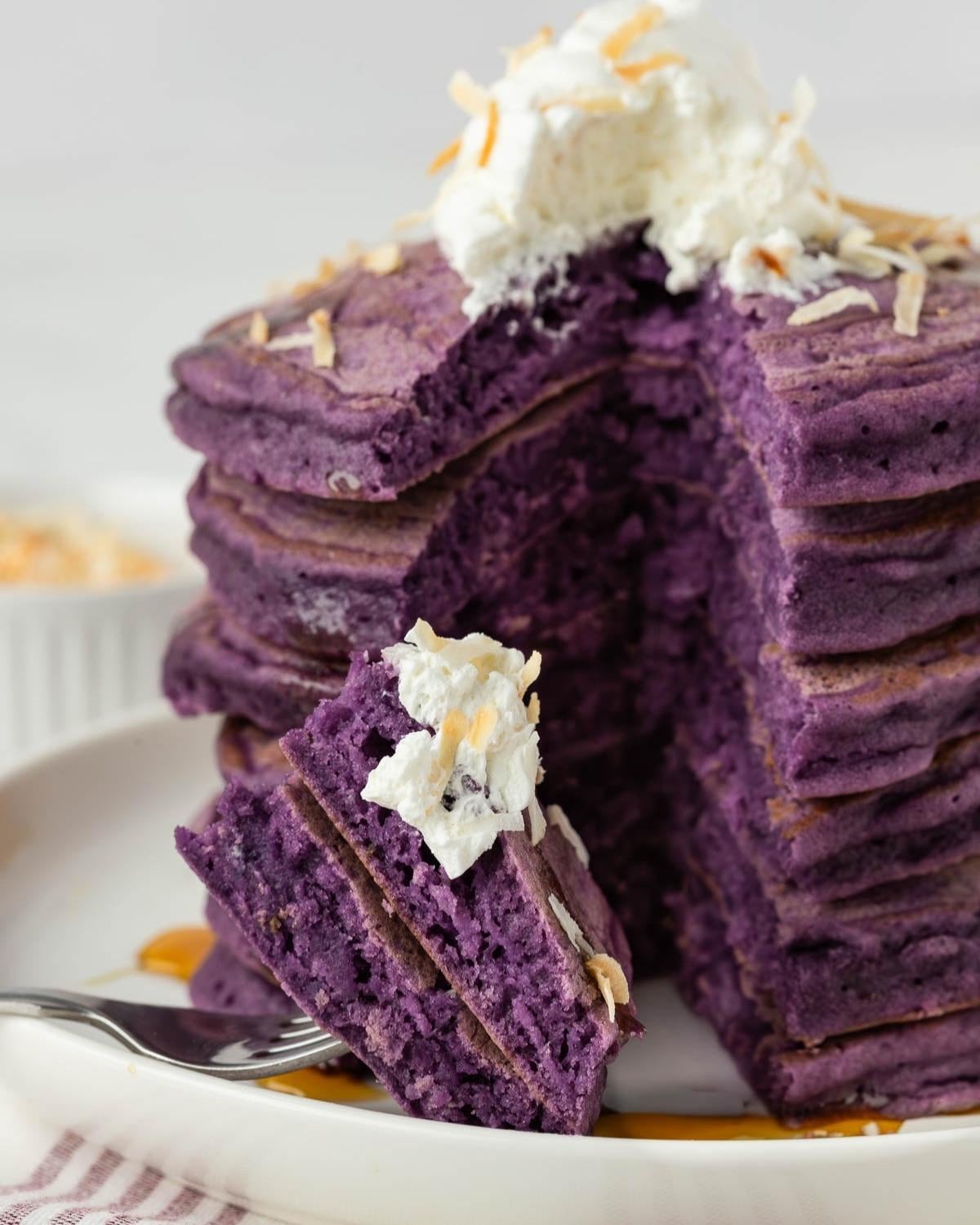 stack of purple ube pancakes on a plate, with a piece on a fork to show their fluffy texture. They're topped with coconut whipped cream and toasted shredded coconut