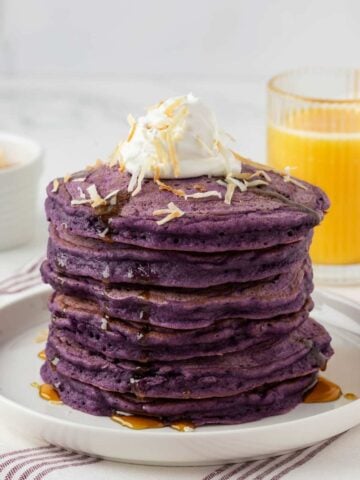 stack of purple ube pancakes on a plate topped with coconut whipped cream and toasted shredded coconut