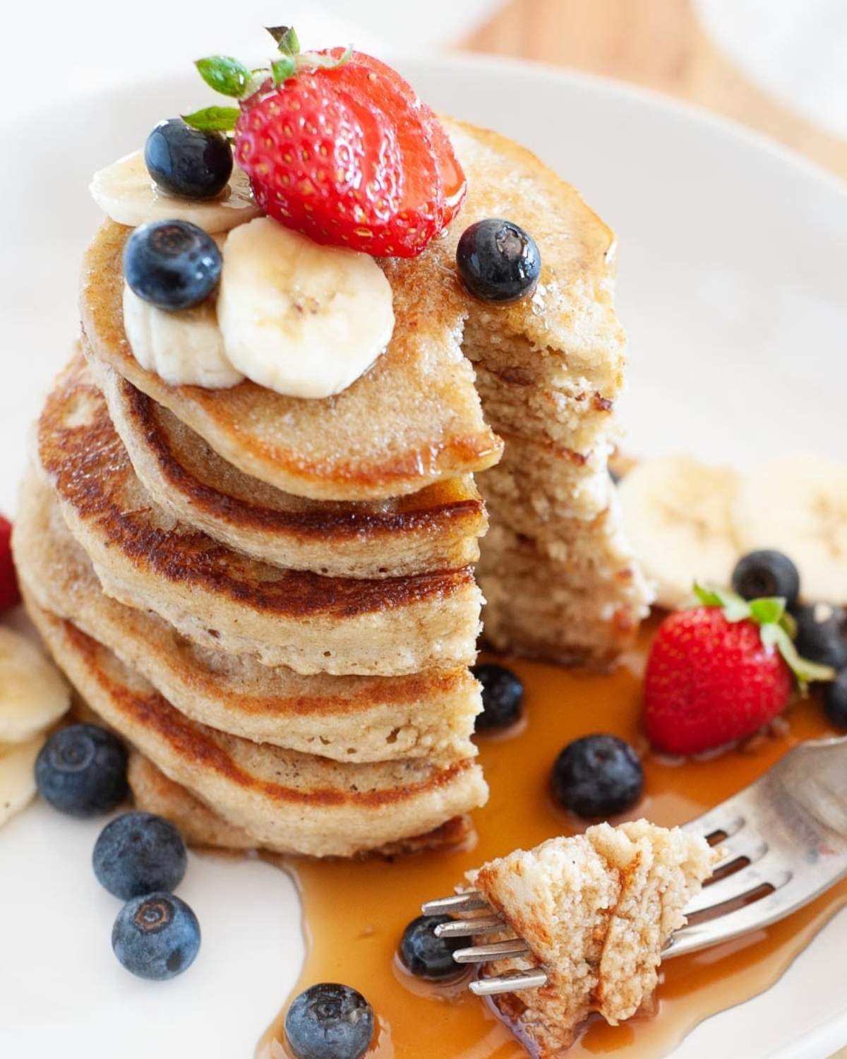 stack of healthy pancakes made with oats on a plate sliced to show fluffy texture. They're topped with fresh berries and banana slices with a drizzle of syrup