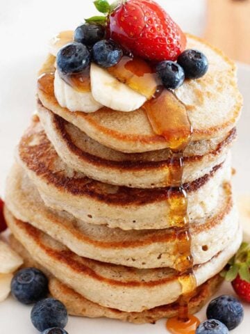stack of healthy pancakes made with oats on a plate topped with fresh berries and banana slices with a drizzle of syrup