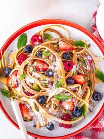 overhead view of pancake batter cooked to look like spaghetti strands on a plate, with fresh fruit and powdered sugar on top as toppings