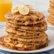 fluffy stack of peanut butter banana pancakes on a plate topped with banana slices, peanut butter drizzle and some syrup
