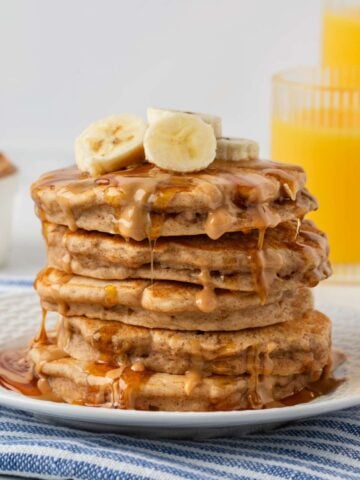 fluffy stack of peanut butter banana pancakes on a plate topped with banana slices, peanut butter drizzle and some syrup