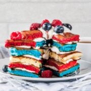 stack of patriotic pancakes of red white and blue colors on a plate. Topped with berries and whipped topping, sliced to show their fluffy texture