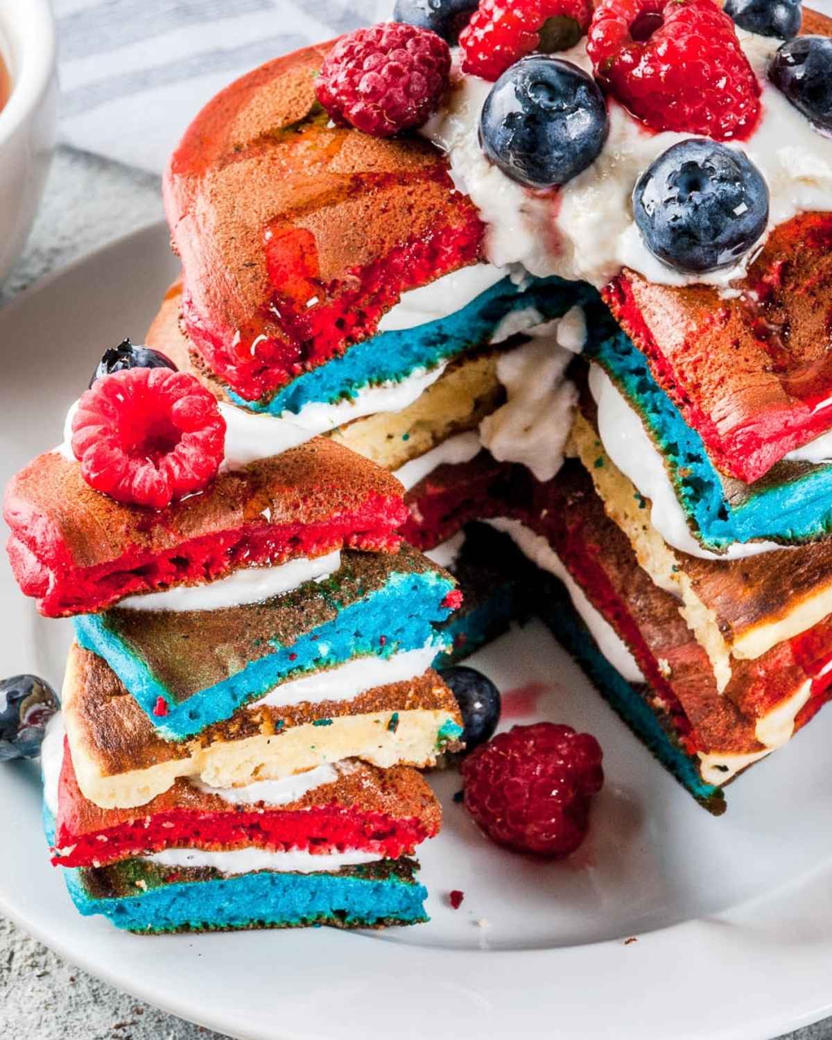 stack of patriotic pancakes of red white and blue colors on a plate. Topped with berries and whipped topping, sliced to show their fluffy texture