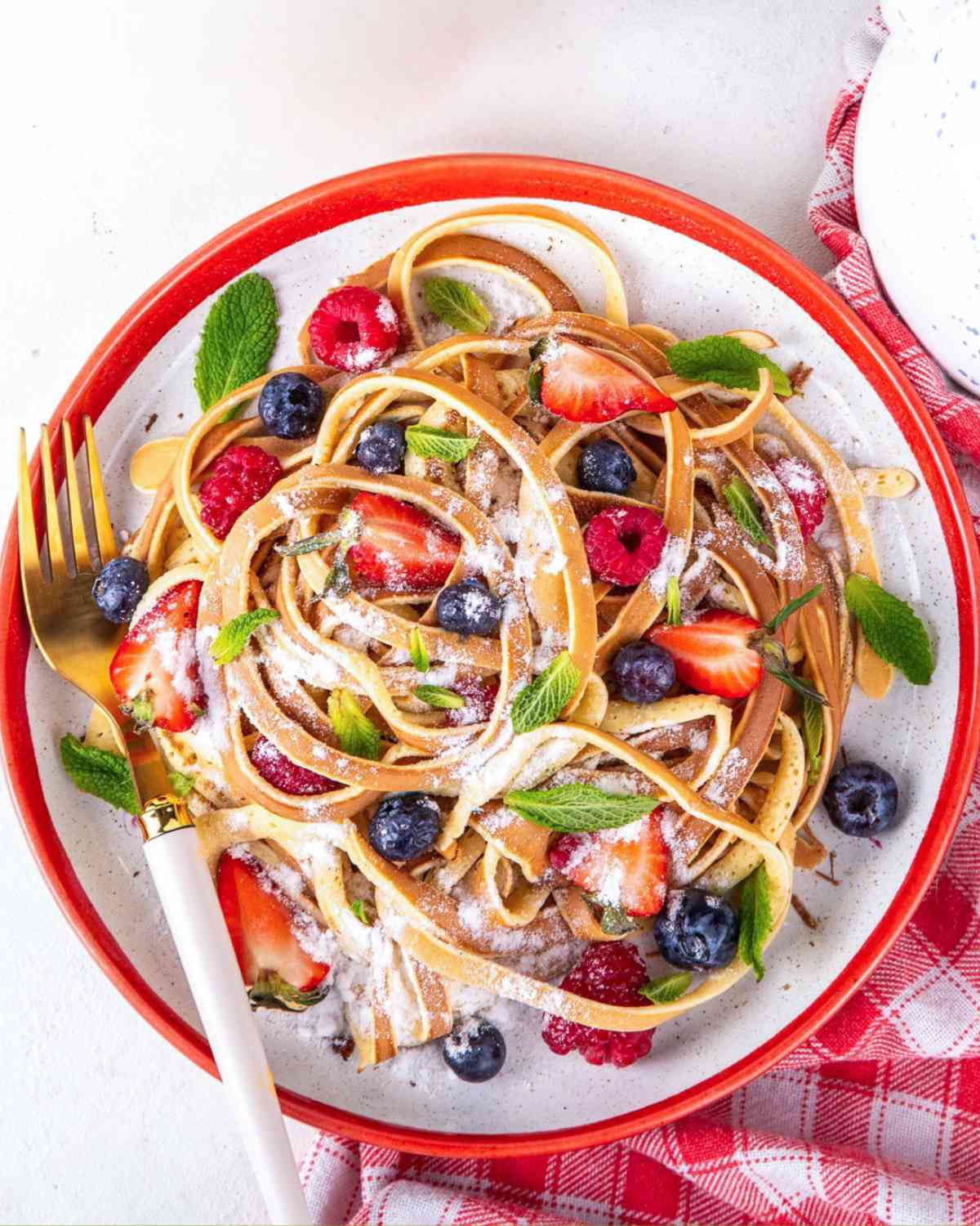 pancake spaghetti on a plate which is pancake batter cooked to look like spaghetti strands on a plate, with fresh fruit and powdered sugar on top as toppings