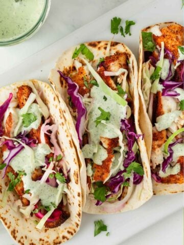 three fish tacos with cilantro sauce and slaw on a white tray