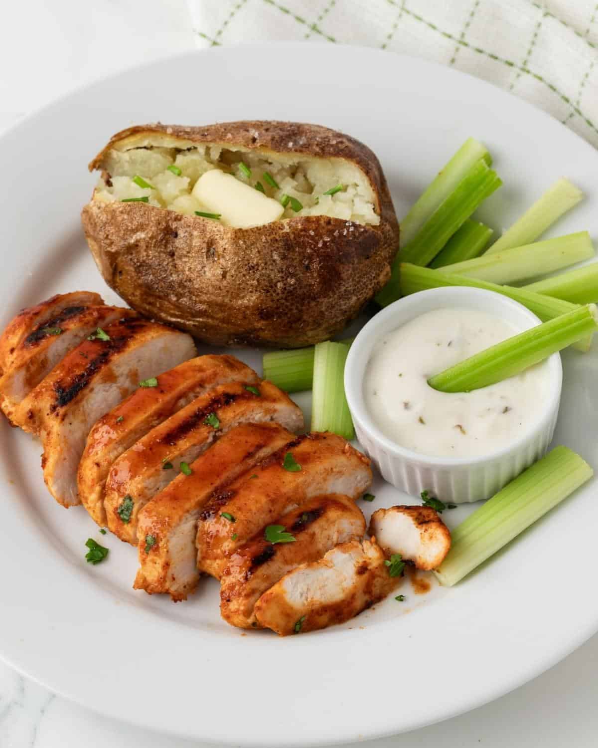 buffalo chicken breasts served on a plate with a baked potato and celery sticks with ranch