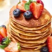 delicious stack of french toast pancakes on a plate topped with diced strawberries and blueberries with syrup. The pancakes are golden brown, dipped in a cinnamon egg custard like french toast.