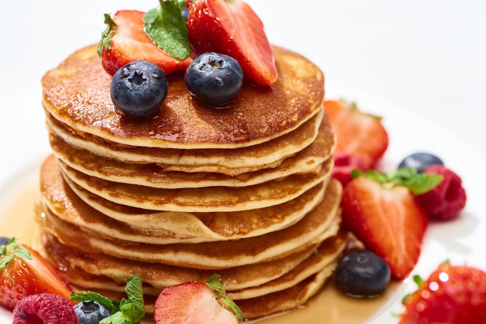 delicious stack of french toast pancakes on a plate topped with diced strawberries and blueberries with syrup. The pancakes are golden brown, dipped in a cinnamon egg custard like french toast.
