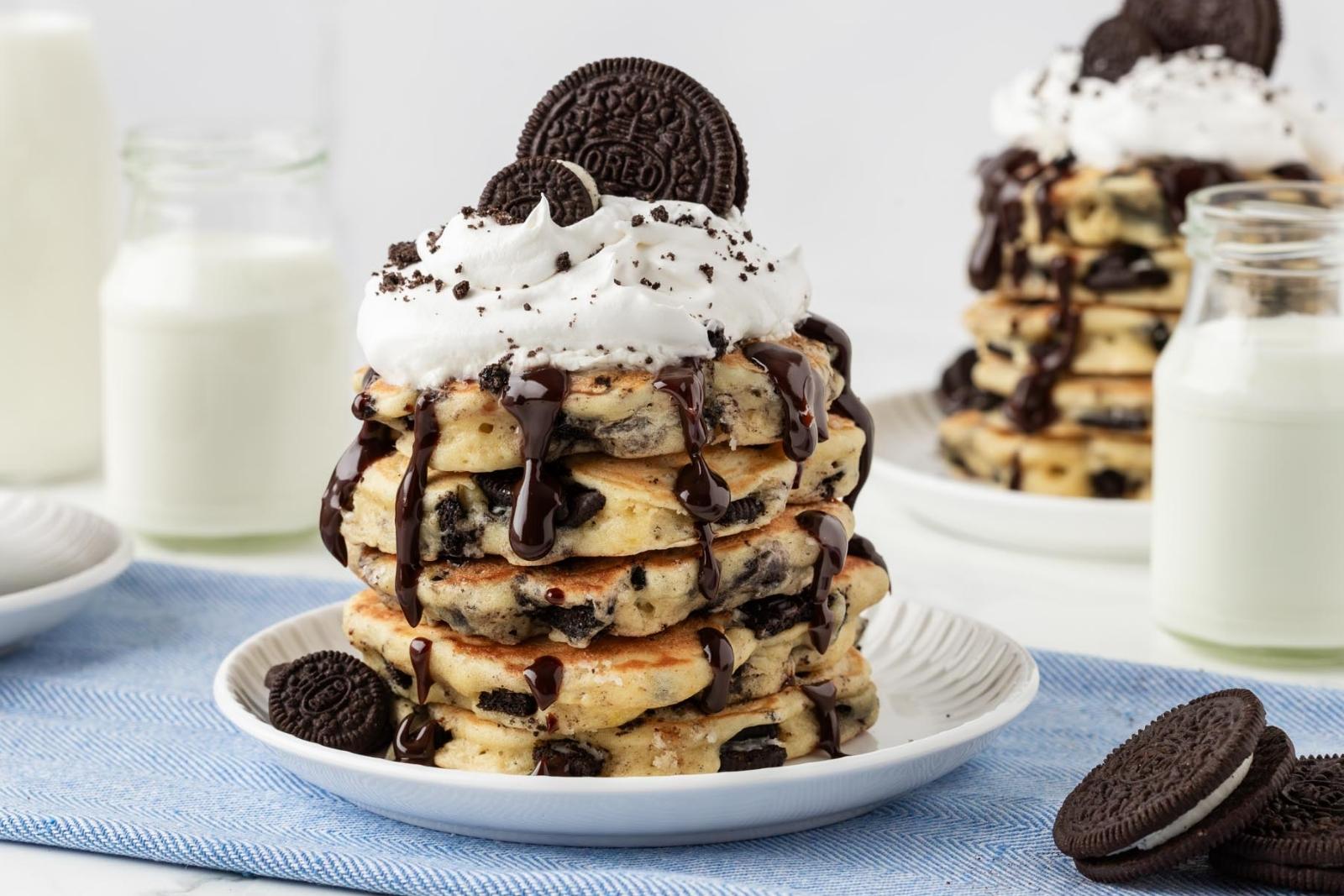 stack of oreo pancakes on a plate topped with chocolate syrup, whipped cream, and oreos on top