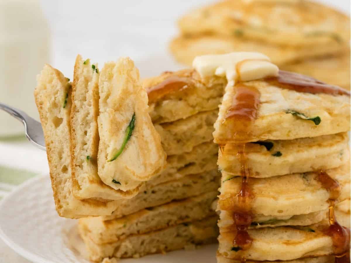 stack of fluffy zucchini pancakes on a plate topped with butter and pancake syrup, sliced to show the fluffy texture poked on a fork