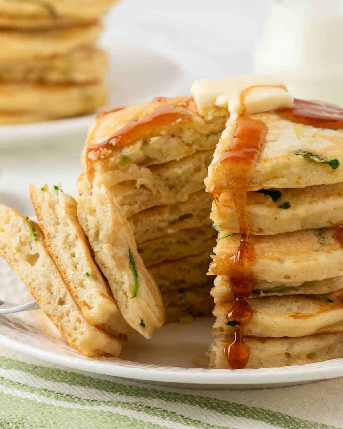stack of fluffy zucchini pancakes on a plate topped with butter and pancake syrup, sliced to show the fluffy texture poked on a fork