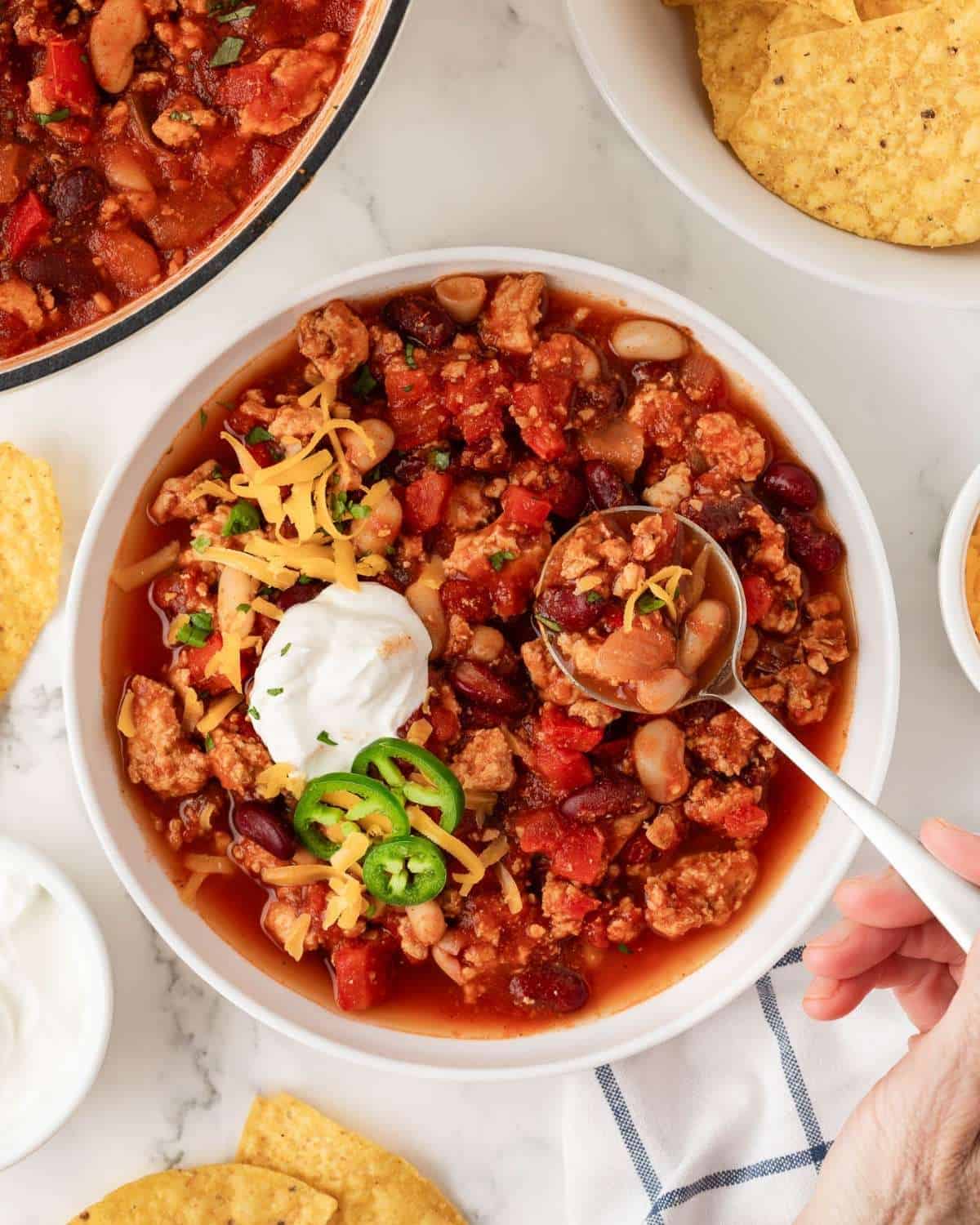 overhead view of a bowl of chicken chili being spooned out of the bowl, topped with sour cream, a little cheese. some chips on the side.