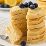 fluffy stack of lemon pancakes on a plate, topped with blueberries and lemon zest. the stack has been sliced and a couple pieces are pierced through a fork to show their fluffy texture