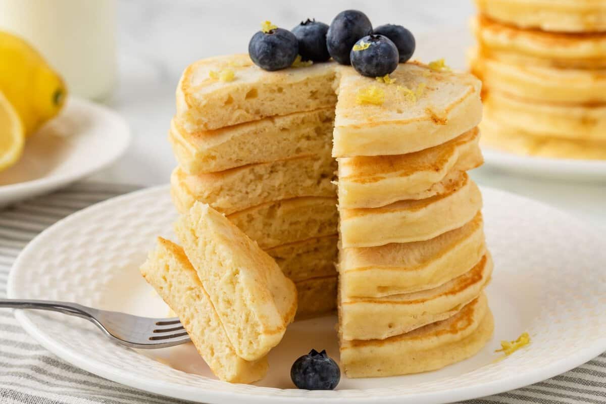 fluffy stack of lemon pancakes on a plate, topped with blueberries and lemon zest. the stack has been sliced and a couple pieces are pierced through a fork to show their fluffy texture