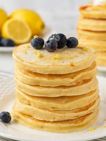 fluffy stack of lemon pancakes on a plate, topped with blueberries and lemon zest.