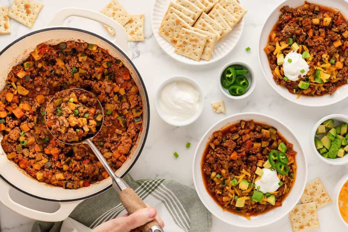 overhead view of a dutch oven with chili with vegetables using a ladle to serve with two bowls on the table filled with chili and topped with avocado, cheese, and sour cream and crackers on the side