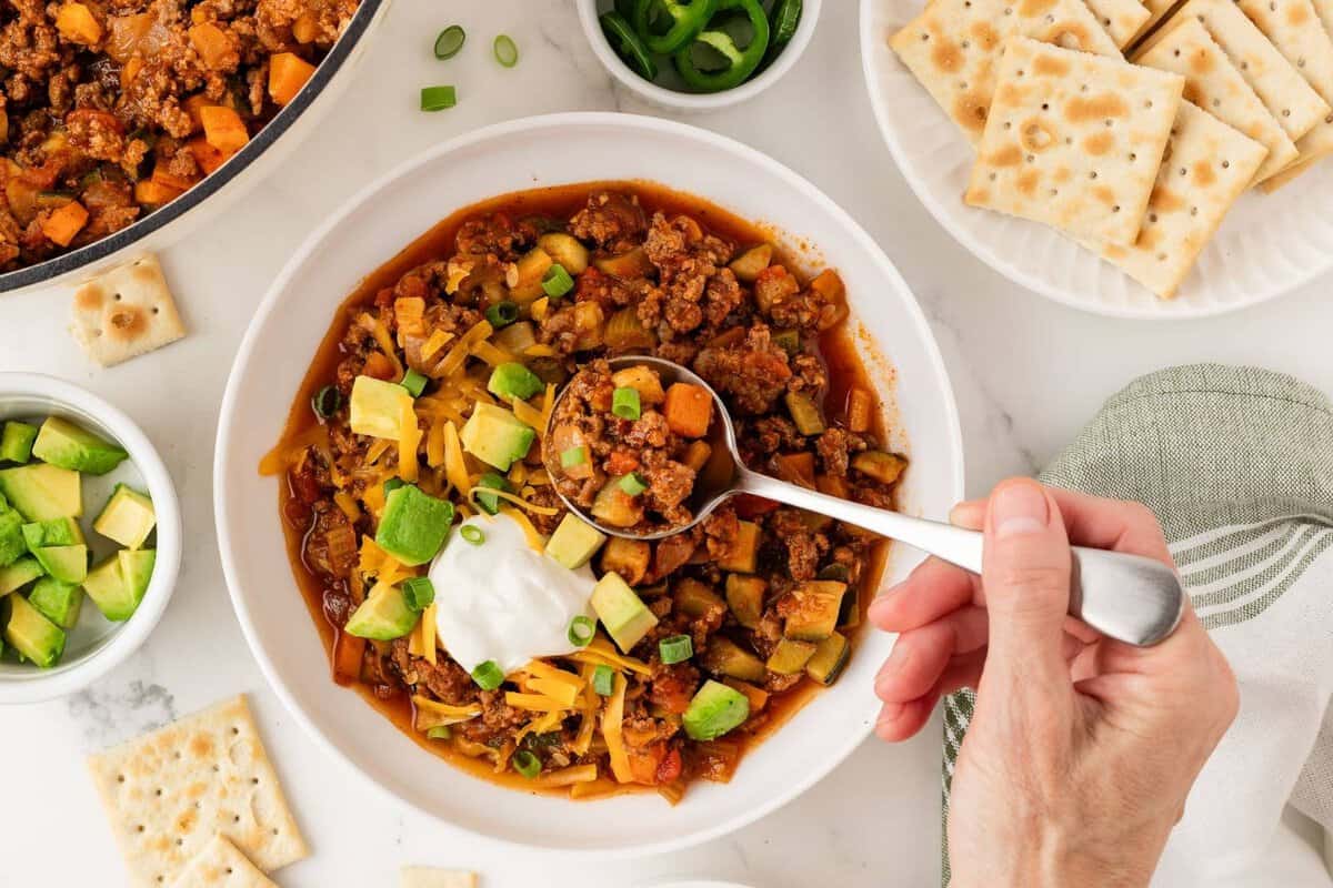 overhead view of a bowl of chili with vegetables topped with avocado, cheese, and some sour cream with a hand spooning out some