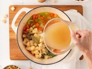 a hand pouring broth in a pot with veggies and potatoes